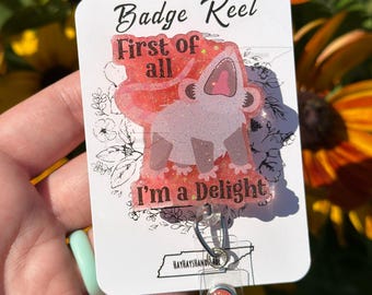 May include: A white badge reel with the text "Badge Reel First of all I'm a Delight" and a cartoon character. The reel has a clear plastic strap and a metal clip. The background is blurred with yellow and orange flowers.