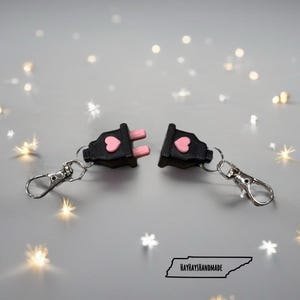 May include: Two black plug-shaped keychains with pink prongs and a pink heart. Each keychain has a silver clasp. The keychains are separated, with the text "HAYHAYSHANDMADE" and a Tennessee outline in the background.