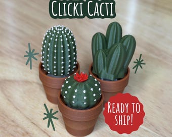Może przedstawiać: Trzy małe kaktusy w doniczkach, jeden z czerwonym kwiatem na górze, z tekstem "Clicki Cacti" i "Ready to Ship!" na drewnianej powierzchni.