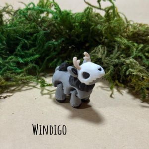 May include: A 3D-printed figurine of a Windigo, featuring a white skull-like head with antlers, a black and gray body, and four legs. The figurine is set against a backdrop of green moss and a tan surface. The word "WINDIGO" is printed below.