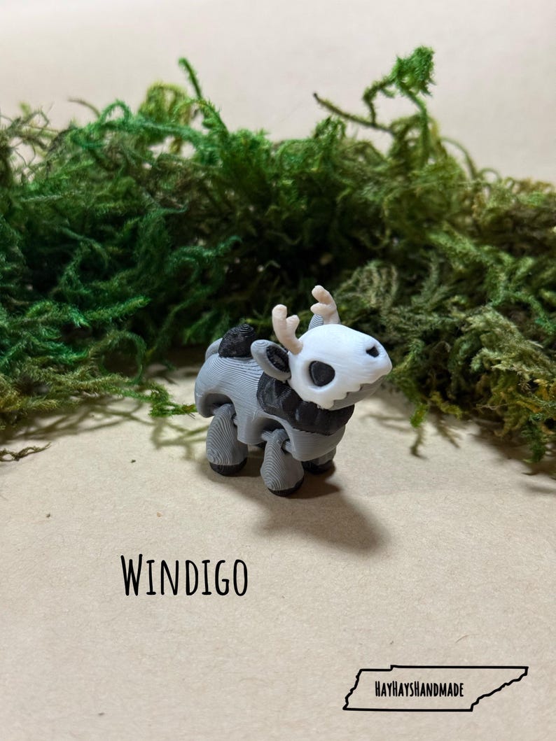 May include: A 3D-printed figurine of a Windigo, featuring a white skull-like head with antlers, a black and gray body, and four legs. The figurine is set against a backdrop of green moss and a tan surface. The word "WINDIGO" is printed below.