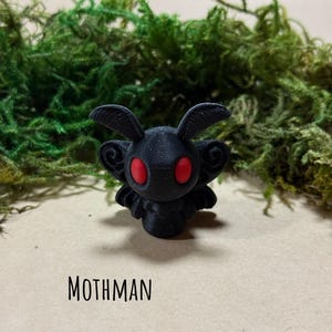 May include: A black Mothman figurine with red eyes, set against a backdrop of green moss. The word "MOTHMAN" is printed below the figurine, with "HAYHAYSHANDMADE" and a Tennessee outline at the bottom.