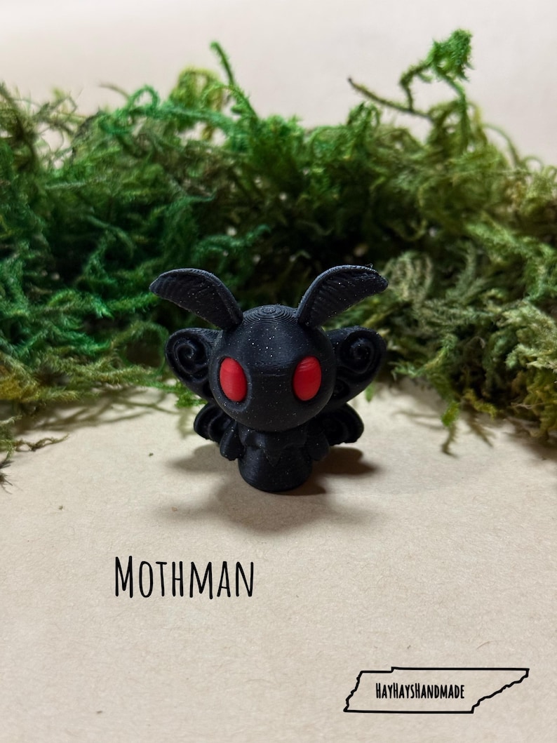 May include: A black Mothman figurine with red eyes, set against a backdrop of green moss. The word "MOTHMAN" is printed below the figurine, with "HAYHAYSHANDMADE" and a Tennessee outline at the bottom.