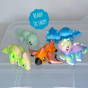 May include: A collection of colorful dinosaur figurines in various shades of green, blue, orange, and purple. Each figurine has a unique design and is set against a clear plastic container. The text "READY TO SHIP!" is on a blue circle.