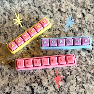 May include: Three personalized keychains with names: "AUTUMN" in pink on yellow, "AURORA" in pink on blue, and "HALEY" in pink with a heart on pink. Each keychain has a silver clasp.