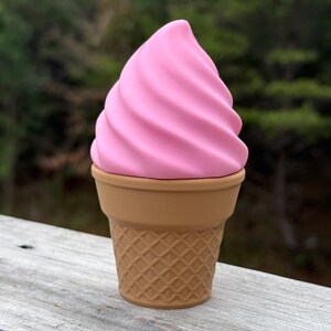 May include: A whimsical ice cream-shaped container with a pink swirl top and a brown cone base. The container is made to look like a soft-serve ice cream cone. The base has a waffle cone pattern.