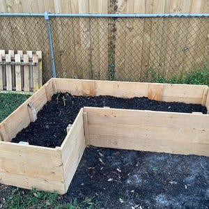May include: A wooden raised garden bed in the shape of an L. The bed is filled with dark brown soil and has a few small green plants growing in it.