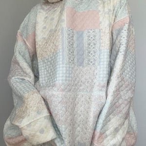 May include: A pastel patchwork hoodie with a drawstring hood and kangaroo pocket. The design incorporates floral, striped, and patterned fabrics. The quilted details and varied textures create a unique, cosy aesthetic. The hoodie is made of soft fabric.