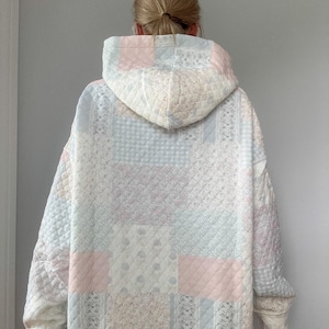 May include: A quilted patchwork hoodie in pastel colors, including pink, blue, and cream. The hoodie features a variety of fabric patterns and textures, with a quilted design throughout. The sleeves and hood are lined with a soft fabric.