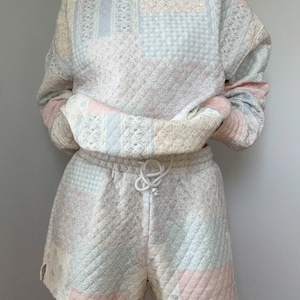 May include: A pastel patchwork sweatshirt and shorts set. The crew neck top has long sleeves, and the shorts have a drawstring waist. The fabric features a mix of floral, striped, and geometric patterns in pink, blue, and cream. The set is quilted.