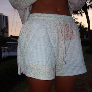 May include: Patchwork quilted shorts with a drawstring waist. The shorts feature sections of white, light blue, and pink floral patterns. A matching jacket is visible above the shorts. The shorts are made of a soft, quilted material.