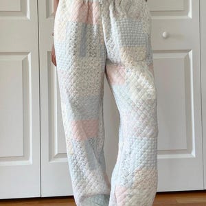 May include: Pastel patchwork sweatpants with a drawstring waist. The trousers are made from quilted fabric and feature a mix of patterns and colours, including pink, blue, and white. The design incorporates various quilted textures and a relaxed fit.