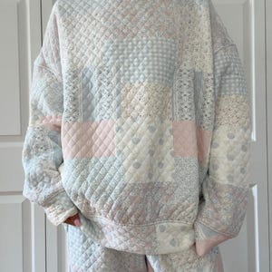 Puede incluir: Conjunto de sudadera y pantalón patchwork acolchado. El conjunto presenta un cuello alto y está confeccionado con varias telas pastel, incluyendo rosa, azul y crema. El diseño incluye patrones florales y geométricos, creando una estética vintage y acogedora.