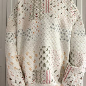 May include: A quilted, oversized sweatshirt in white with a patchwork design featuring Christmas trees, candy canes, gingerbread men, and other holiday motifs. The sweatshirt has a high collar and diamond quilting.