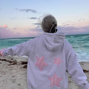Może przedstawiać: Jasnoszara bluza z kapturem z trzema różowymi aplikacjami w kształcie gwiazd na plecach. Bluza ma kaptur i różowe mankiety. Osoba stoi na piaszczystej plaży z oceanem w tle.