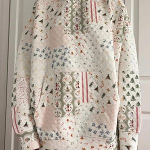 Może przedstawiać: Biały pikowany zestaw z kapturem i szortami z patchworkowym wzorem. Wzór zawiera choinki, piernikowe ludziki, laski cukrowe i inne elementy o tematyce świątecznej. Bluza z kapturem ma przednią kieszeń i kaptur.