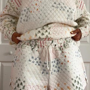 May include: A quilted two-piece set featuring a sweatshirt and shorts. The set is white with a patchwork design of Christmas trees, gingerbread men, and candy canes. The shorts have a drawstring waist.