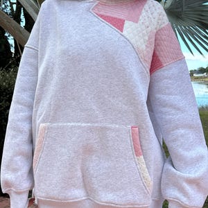 May include: A light gray heathered hoodie featuring a quilted patchwork panel on the upper right chest and shoulder in shades of pink and white. The garment includes a large front kangaroo pocket with matching pink and white quilted fabric accents.
