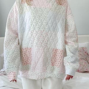 Op de afbeelding: Een gewatteerde patchwork-sweatshirt in pastelkleuren. Het ontwerp heeft vierkanten van roze, witte en lichtblauwe stof met bloemen- en streepjespatronen. De sweatshirt heeft een hoge halslijn en lange mouwen. De persoon draagt een witte joggingbroek.