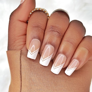 Könnte beinhalten: Nahaufnahme einer Hand mit quadratischen, weißen und klaren Acrylnägeln. Jeder Nagel hat eine weiße Spitze mit einem goldenen Chevron-Design. Die Hand trägt einen goldenen Ring und ruht auf einem braunen Stoff.