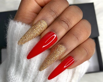 May include: A hand displaying long, stiletto-shaped artificial nails. The nails alternate between a vibrant red and a gold glitter finish. The hand is resting on a white, textured surface, with a black box in the background.