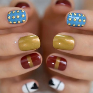 Peut inclure: Gros plan sur une main avec des ongles peints. Les ongles sont peints de différentes couleurs, dont le bleu avec des clous dorés, l'or, le bordeaux avec une bande dorée et le blanc avec un motif triangulaire noir. Les ongles sont carrés.