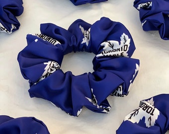 May include: Several blue scrunchies with a white maple leaf design and the text "TORONTO MAPLE". The scrunchies are made of a silky material and are scattered on a white surface.