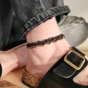 May include: A black ankle bracelet with gold beads. The bracelet is made of a delicate, intricate design.