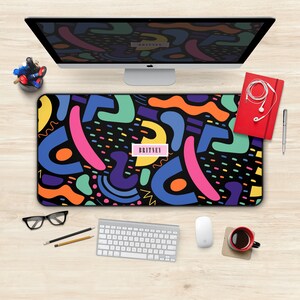 May include: A black desk mat with a colorful abstract pattern featuring geometric shapes and dots. The mat has the name "BRITNEY" printed in pink letters.