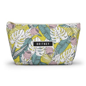 Puede incluir: Un neceser con cremallera con un estampado de hojas y flores tropicales en amarillo, blanco, turquesa y rosa. La bolsa tiene el nombre "BRITNEY" impreso en un rectángulo blanco.