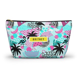 May include: A zippered cosmetic bag with a vibrant tropical pattern. The design features flamingos, palm trees, bananas, and watermelon slices in shades of pink, black, and turquoise. The name "BRITNEY" is displayed on a yellow rectangle.