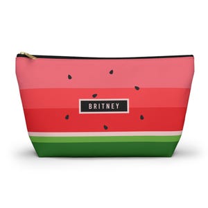 May include: A watermelon-themed cosmetic bag with a zipper closure. The bag features a pink and red striped design, black seed accents, and the name "BRITNEY" in a white rectangle. The bottom of the bag is green.