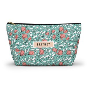 May include: A teal cosmetic bag with a floral pattern of pink flowers and white leaf-like shapes. The bag has a black zipper and a rectangular label that reads "BRITNEY".