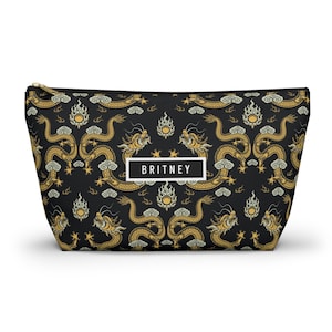 May include: Black cosmetic bag with a gold dragon pattern and the name "BRITNEY" in a white rectangle. The bag has a gold zip and a repeating pattern of dragons, clouds, and suns.