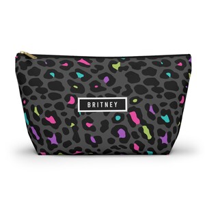 Peut inclure: Une trousse de maquillage zippée avec un imprimé léopard gris et des accents colorés en rose, turquoise, violet et vert citron. Le sac porte le nom "BRITNEY" dans un rectangle blanc.