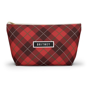 May include: A red plaid cosmetic bag with a gold zipper. The bag features a repeating pattern of red, brown, and white lines. The name "BRITNEY" is printed in a white rectangle on the front of the bag.