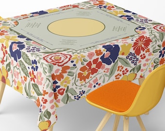 May include: A floral tablecloth with a cream background and a colorful pattern of red, yellow, blue, and pink flowers. The tablecloth is on a wooden table with a yellow chair with an orange seat. The tablecloth has a central design with text.