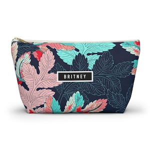 Può includere: Un beauty case blu navy con un motivo floreale colorato con foglie rosa, turchese e rosse. La borsa ha una cerniera dorata e il nome "BRITNEY" in una scatola bianca.