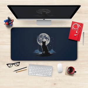 Pode incluir: Um tapete de mesa azul com uma silhueta de gato preto olhando para cima para uma lua cheia. A lua é branca com uma superfície texturizada. O gato está sentado em uma pequena rocha.