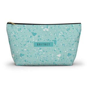 Puede incluir: Un neceser turquesa con un estampado de terrazo de formas blancas y turquesas. La bolsa tiene una cremallera negra y un tirador dorado. El nombre "BRITNEY" está impreso en un recuadro en la parte delantera.