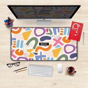 May include: A colorful abstract design desk mat with the word "BRITVEY" in the center. The mat is on a wooden desk with a computer, keyboard, mouse, coffee cup, pencils, and a notebook.