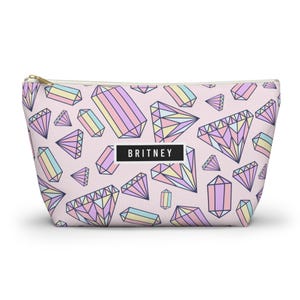 May include: A pink cosmetic bag with a repeating pattern of pastel-colored geometric diamond shapes. The bag has a gold zipper and a black rectangle with the name "BRITNEY" printed in white.