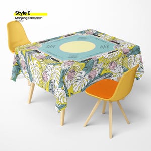 May include: A Mahjong tablecloth with a tropical leaf and flower pattern in yellow, white, pink, and teal. The center of the tablecloth is light blue with a yellow circle. Two yellow chairs with wooden legs and an orange seat are on either side of the table.