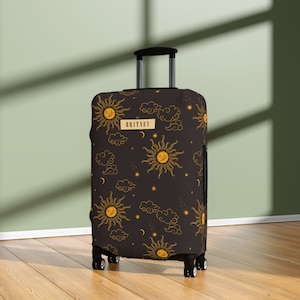 May include: A black suitcase cover featuring a repeating pattern of suns, clouds, stars, and moons in gold. The word "BRITNEY" is displayed on a rectangular gold label. The luggage cover is on a rolling suitcase.