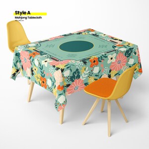 May include: A floral Mahjong tablecloth, 140 x 140 cm, covers a table. The tablecloth features a teal background with a floral pattern in pink, orange, and white. Two yellow chairs with wooden legs and an orange seat are on either side of the table.