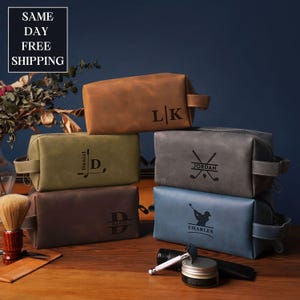 Könnte beinhalten: Sechs Leder-Kulturbeutel in verschiedenen Farben, gestapelt. Farben sind Braun, Grün, Grau und Blau. Jede Tasche ist mit Initialen, Namen oder Golfmotiven personalisiert. Die Taschen sind rechteckig mit Reißverschluss und seitlichem Griff.