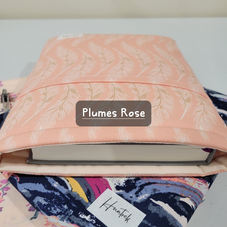 Op de afbeelding: Een roze boekomslag met een wit verenpatroon en de tekst "Plumes Rose". De omslag is gemaakt van stof en heeft een zachte, gestikte textuur.