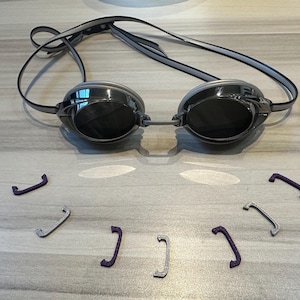 May include: Swimming goggles with dark lenses and a black strap. Several replacement nose bridge pieces in silver and purple are scattered below the goggles. The goggles are designed for competitive swimming and water sports.