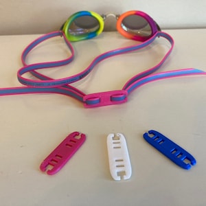May include: A pair of pink and gray swim goggles with a rainbow pattern on the lenses. Three replacement straps are included: pink, white, and blue.