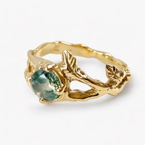 Può includere: Un anello in oro con una pietra preziosa di agata muschiata rotonda e sfaccettata con inclusioni verdi. La fascia è progettata per assomigliare a un ramo d'albero strutturato con piccoli dettagli a foglia in rilievo lungo i lati.